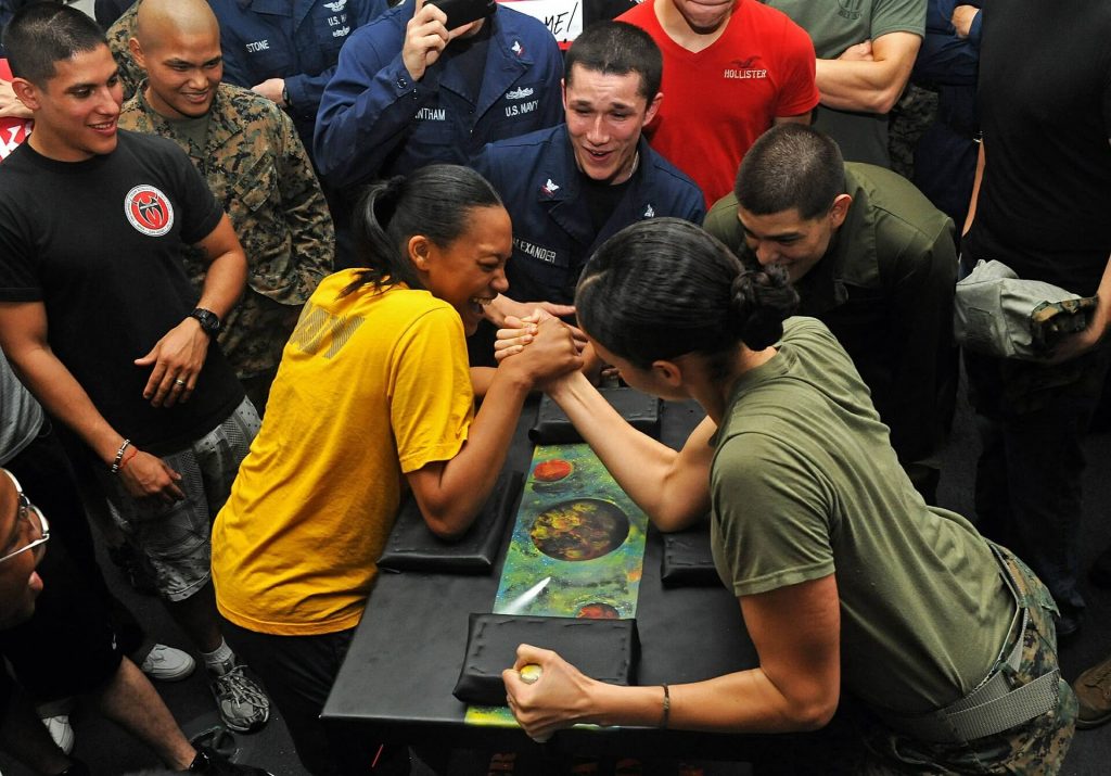 Duas mulheres se enfrentam em uma disputa de queda de braço, enquanto pessoas ao fundo sorriem se divertindo com a competição.